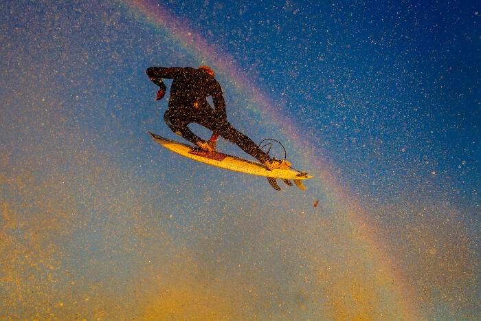 Surfer in a wetsuit airborne above waves with a rainbow in the background, captured in stunning ocean photography.