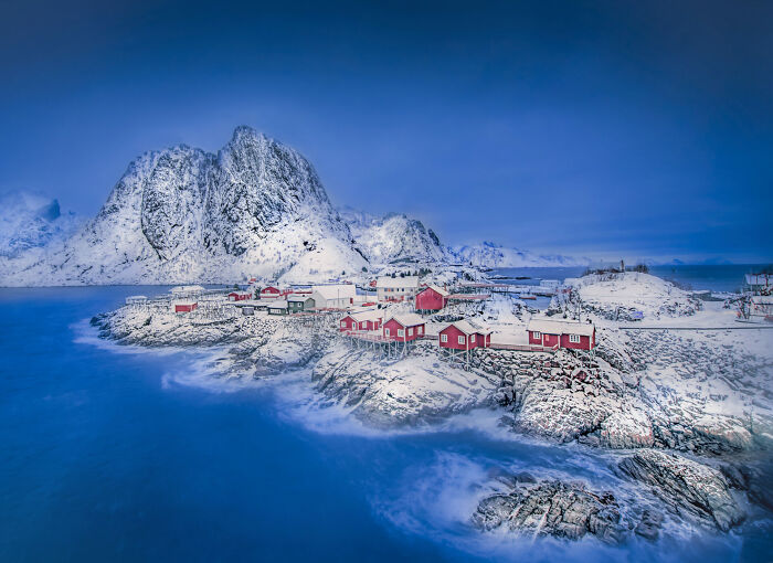 Snow-covered coastal village with red houses and rugged mountains under a moody sky, ocean photography from the 2025 awards.