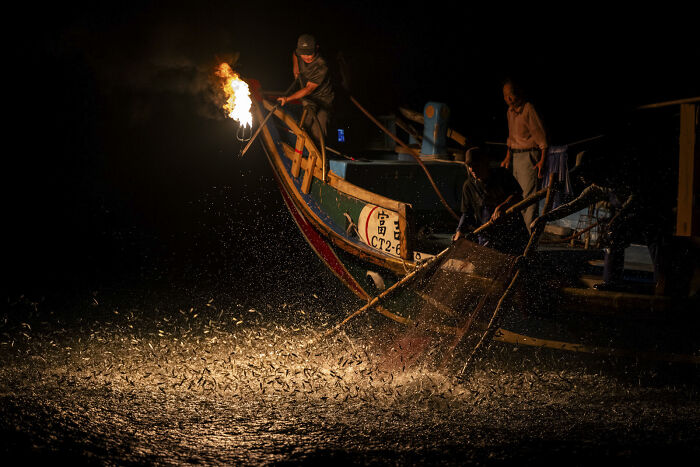 Fishermen using fire on a boat at night to catch fish, captured in a stunning ocean photographer of the year image.