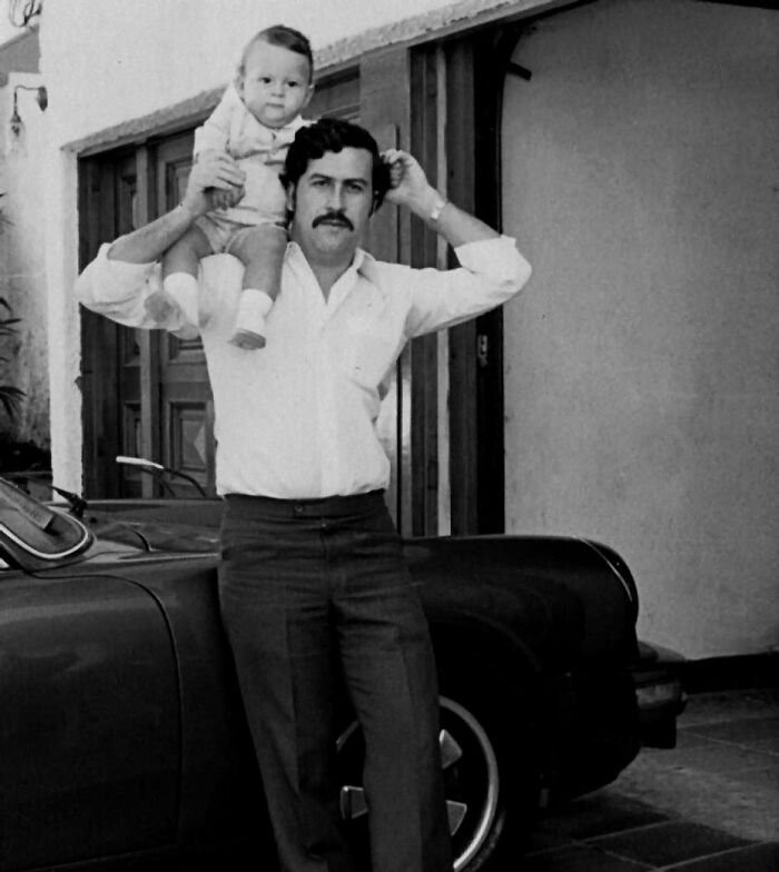 Black and white photo of a notorious mob boss holding a child on his shoulders, standing beside a classic car.