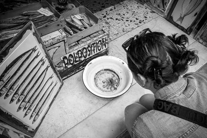 Black and white photograph capturing the hidden poetry of everyday life with a woman observing an octopus in a market setting.