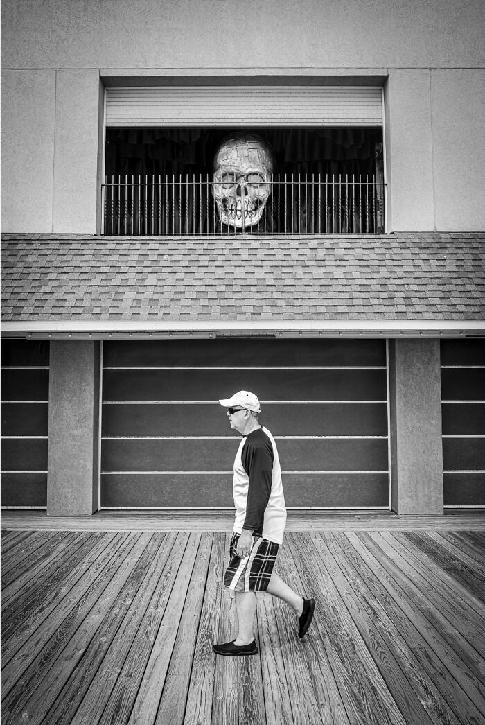 Black and white photograph capturing the hidden poetry of everyday life with a man walking beneath a large skull sculpture.