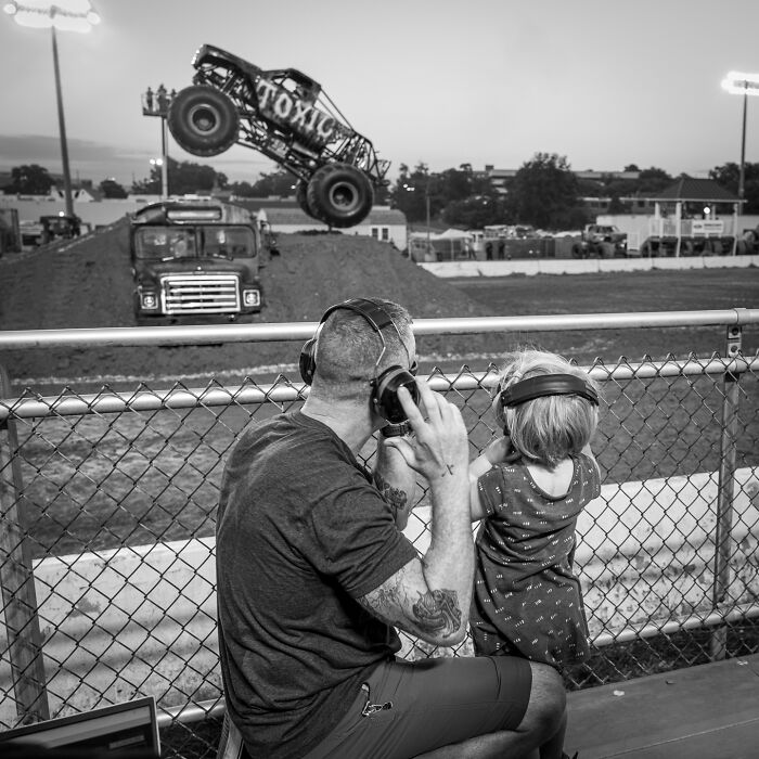 Father and child wearing headphones watching a monster truck jump at an outdoor event, capturing everyday life poetry.