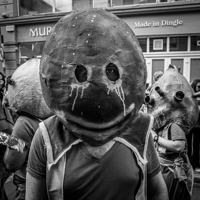 Black and white photograph capturing the hidden poetry of everyday life with people wearing large, artistic masks on a city street.