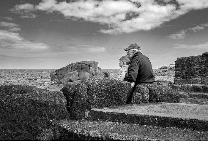 Elderly man and child sitting on rocks by the sea, a striking photograph capturing the hidden poetry of everyday life.