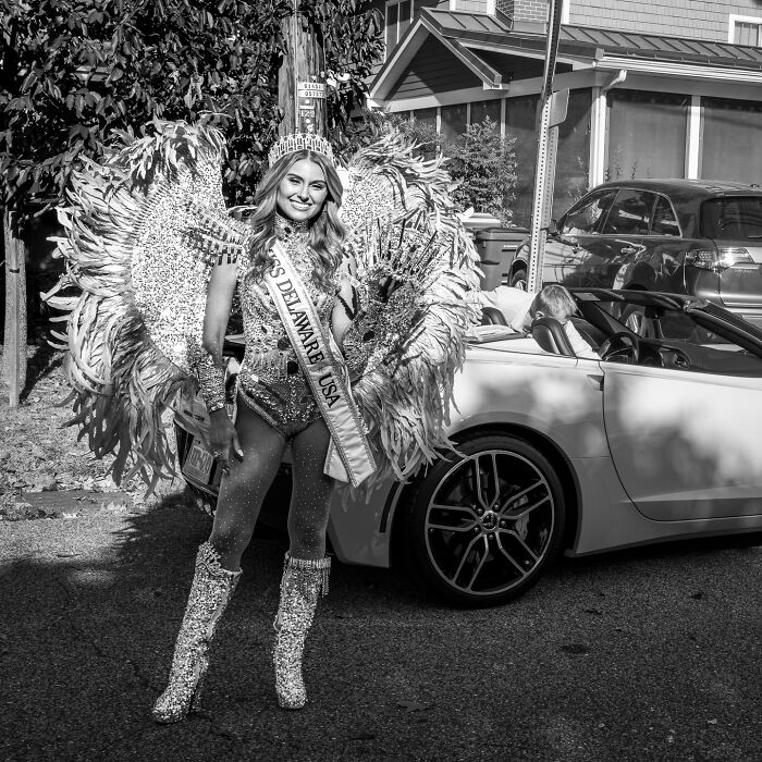 Young woman in an elaborate feathered costume and sash standing beside a convertible car, capturing everyday life poetry.
