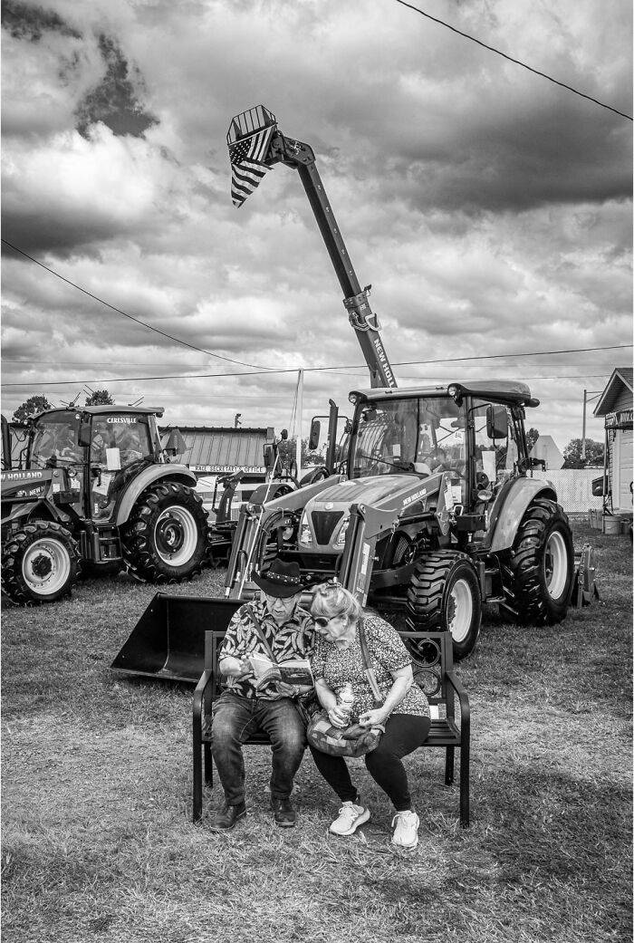 Black and white photograph of two people sitting on a bench near tractors, capturing the hidden poetry of everyday life.