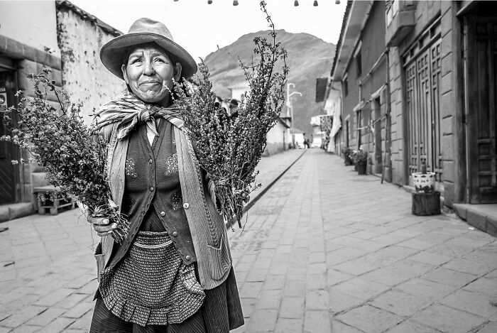 Elderly woman holding bundles of herbs on a quiet street, showcasing the hidden poetry of everyday life in a striking photograph.