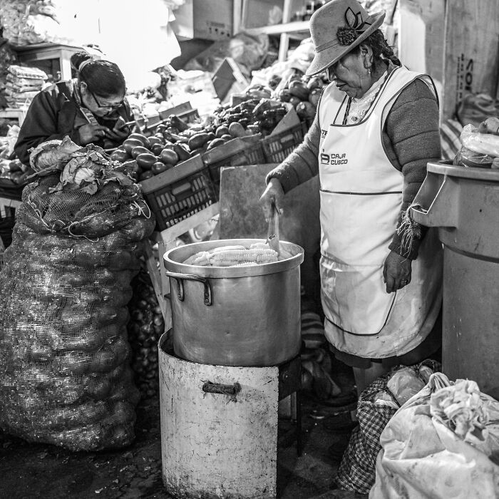 Woman cooking corn at market stall surrounded by vegetables, capturing the hidden poetry of everyday life in black and white.