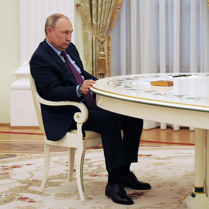 Vladimir Putin sitting at a round table, wearing a dark suit, with focus on his legs amid exoskeleton rumors.