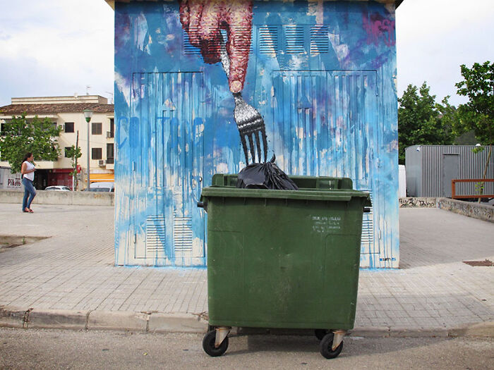 Street art mural of a hand holding a fork above a green dumpster, blending urban environment with creative street art canvas.