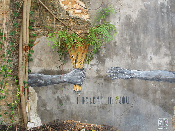 Street art mural showing two hands, one holding plants and the other reaching out, symbolizing connection on a weathered wall.