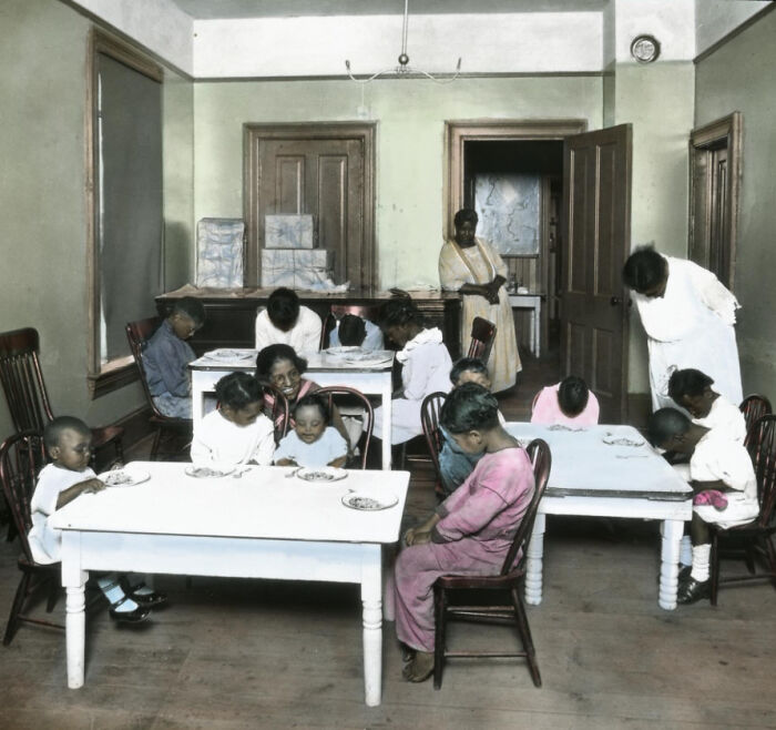 Colorized photo showing African American children and caregivers in a 1920s classroom scene depicting daily life reality.