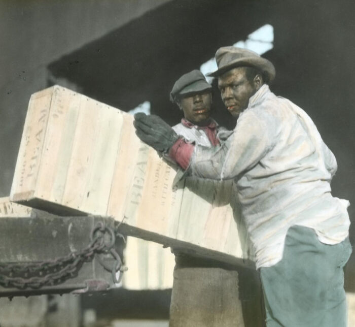 Two African American men unloading a large wooden crate, showing the 1920s work reality in a colorized photo.