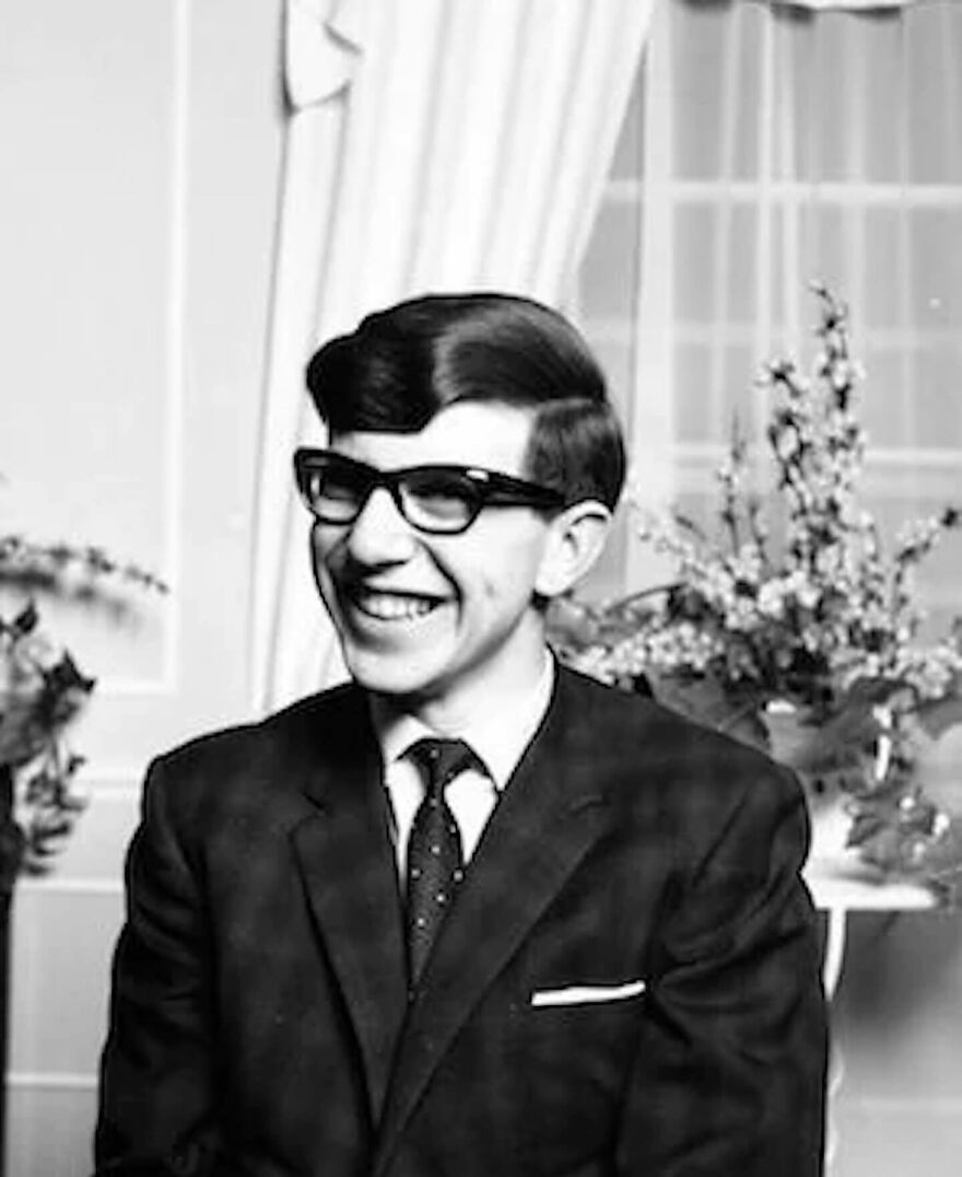 Young Stephen Hawking smiling in a suit and glasses, pictured indoors with floral arrangements behind him, Epstein Island allegations keywords.