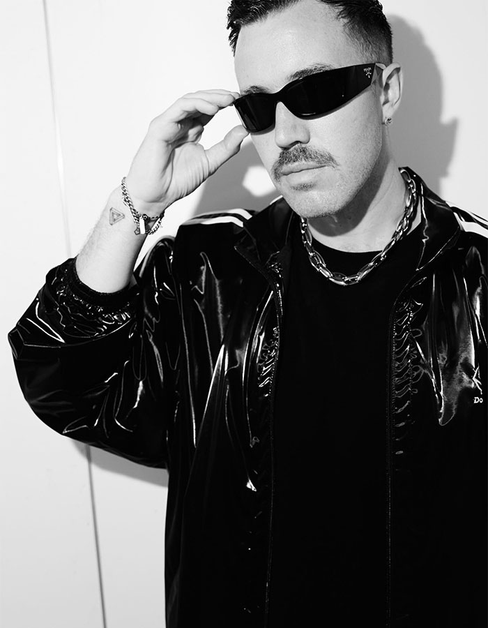 Man wearing sunglasses and a shiny jacket posing for a black and white portrait at an indoor location.