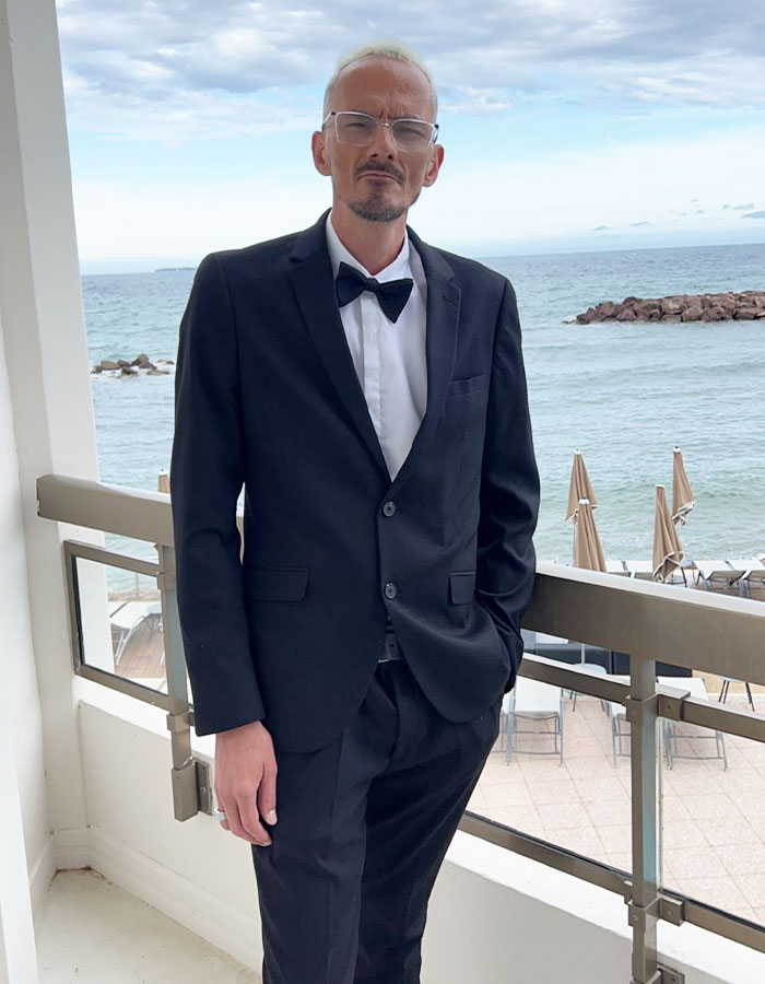 Man in a black suit and bow tie standing on balcony by the sea, related to streamer’s passing and family breaking silence. Man in a black suit and bow tie standing on balcony by the sea, related to streamer’s passing and family breaking silence.
