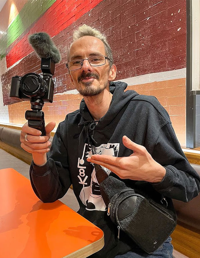 Streamer holding a camera with microphone, wearing glasses and black hoodie, representing streamer’s passing and family reaction. Streamer holding a camera with microphone, wearing glasses and black hoodie, representing streamer’s passing and family reaction.