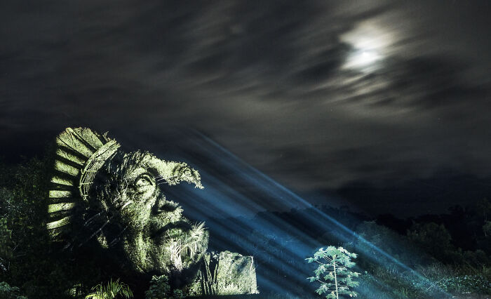 Large street art sculpture of a face illuminated at night with blue light beams under a cloudy moonlit sky.