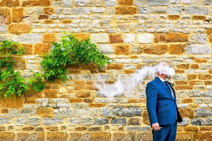 Man in blue suit with cloud of smoke covering his face against a stone wall in a powerful street photo capturing everyday life.