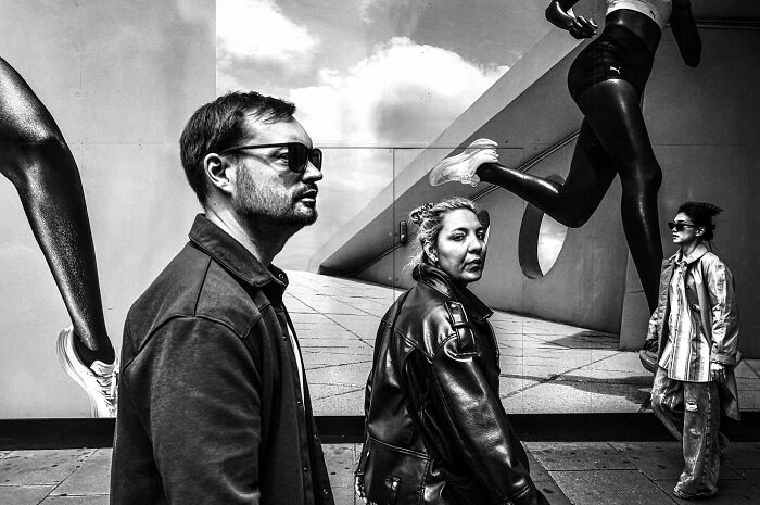 Black and white street photo capturing everyday life with pedestrians and large running statue legs in urban setting.