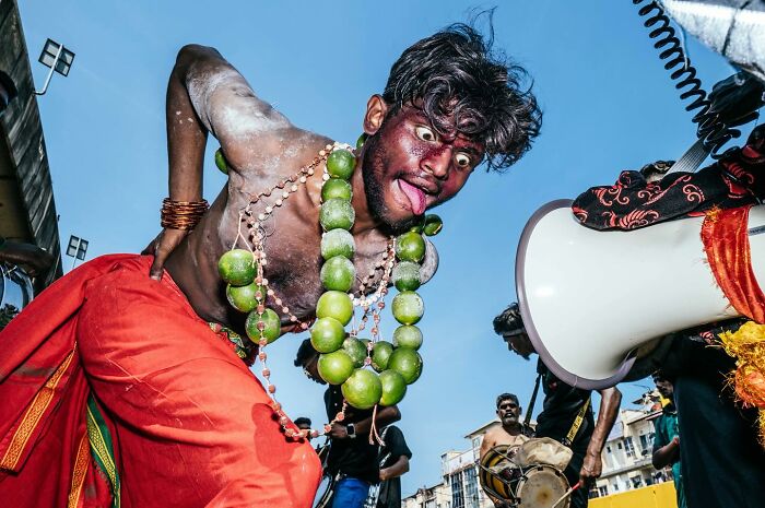 Man with painted face and green bead necklace performing during vibrant street festival, captured in powerful street photo.