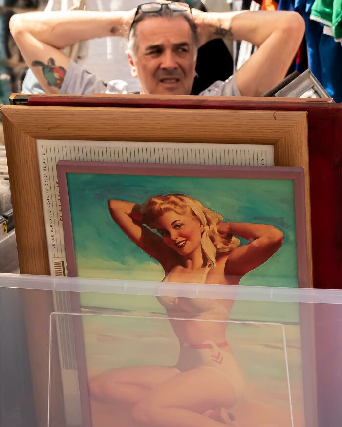 Man with tattoos resting arms behind head behind vintage pin-up art at a street market in a powerful street photo capturing everyday life.