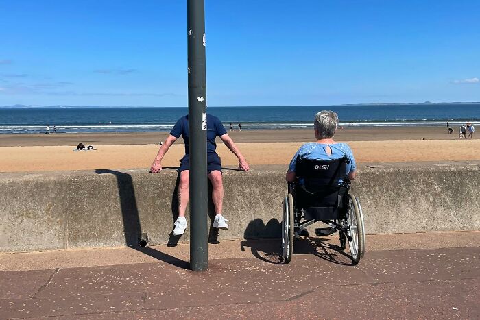 Man sitting on a wall beside a person in a wheelchair with a beach and ocean in the background in a powerful street photo.