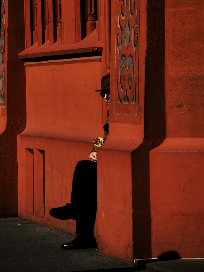 Person in black hat and glasses sitting against a red wall in a powerful street photo capturing everyday life moments.