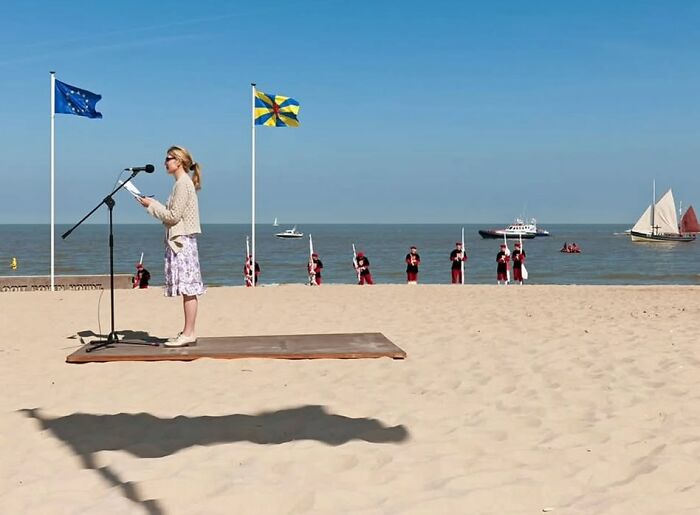 Woman reading at microphone on beach with soldiers standing by water, capturing powerful street photos of everyday life.
