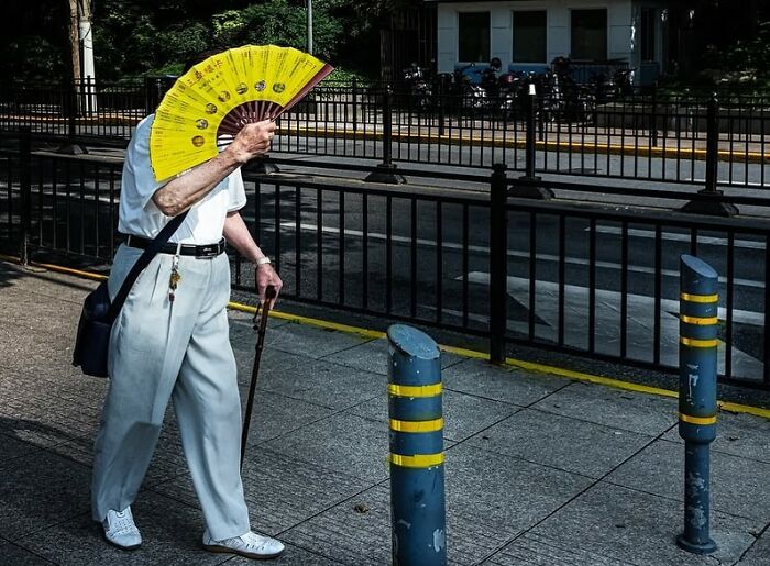 Elderly person walking down the street holding a bright yellow fan, capturing the beauty of everyday life in a street photo.