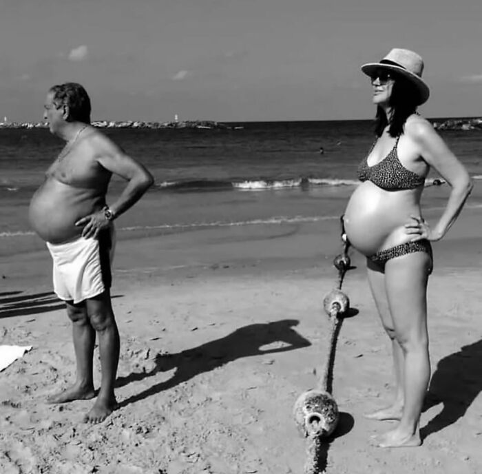 Pregnant man and woman standing on a beach with hands on hips, captured in a powerful street photo of everyday life.