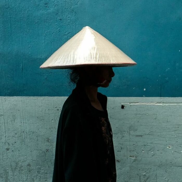Silhouette of person wearing traditional conical hat walking against a textured blue and gray wall in powerful street photos.