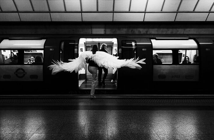 Person wearing large angel wings walking onto a train, a powerful street photo revealing everyday life beauty in black and white.