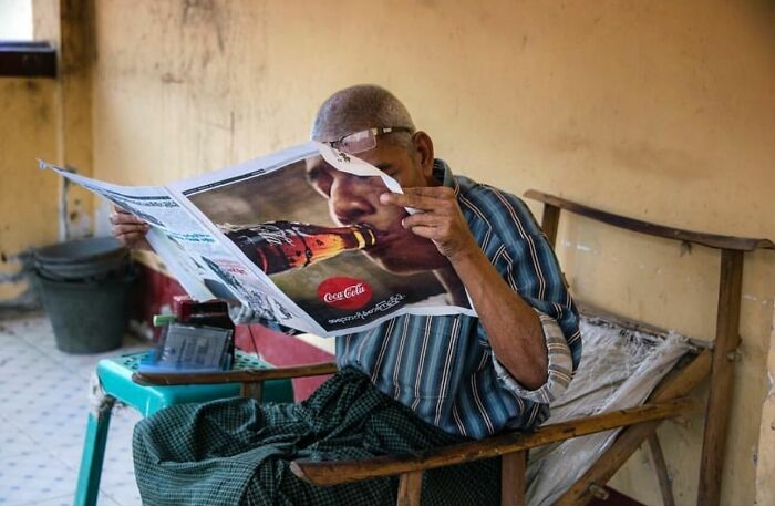Elderly man sitting on a rustic chair reading a newspaper in a simple room, capturing street photos of everyday life.