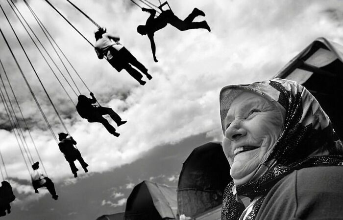 Black and white street photo capturing an elderly woman smiling joyfully near a swinging carousel ride in everyday life.