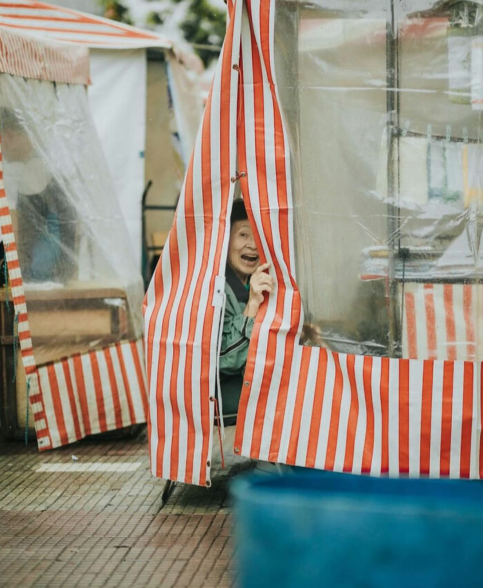 Smiling elderly person peeks through red and white striped fabric on a street, capturing the beauty of everyday life in a powerful moment.