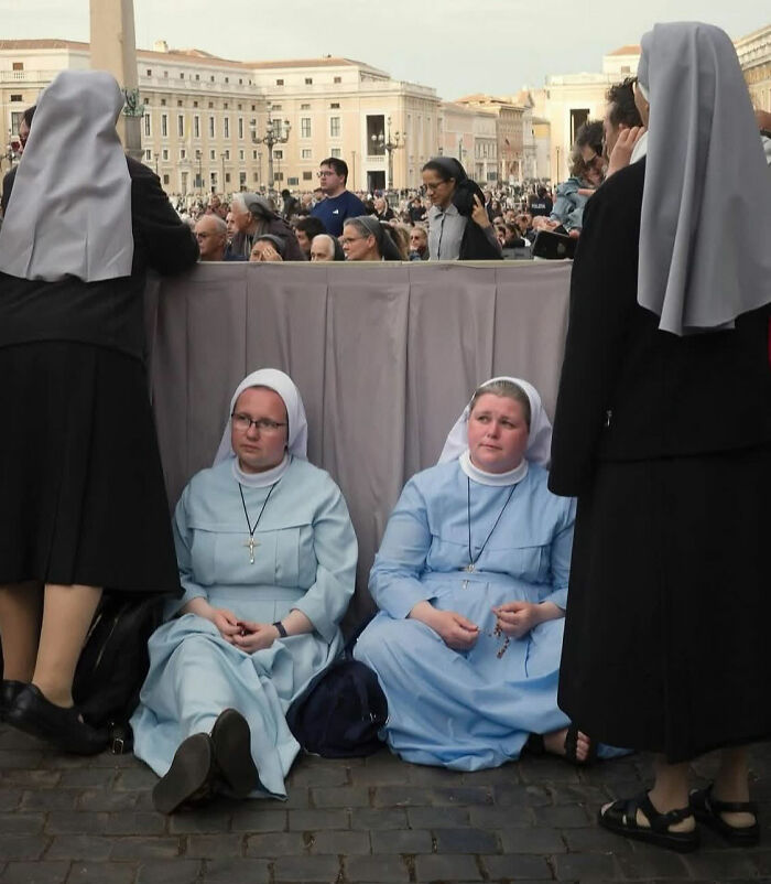 Two nuns sitting on a street curb with a crowd in the background, capturing the beauty of everyday life moments.
