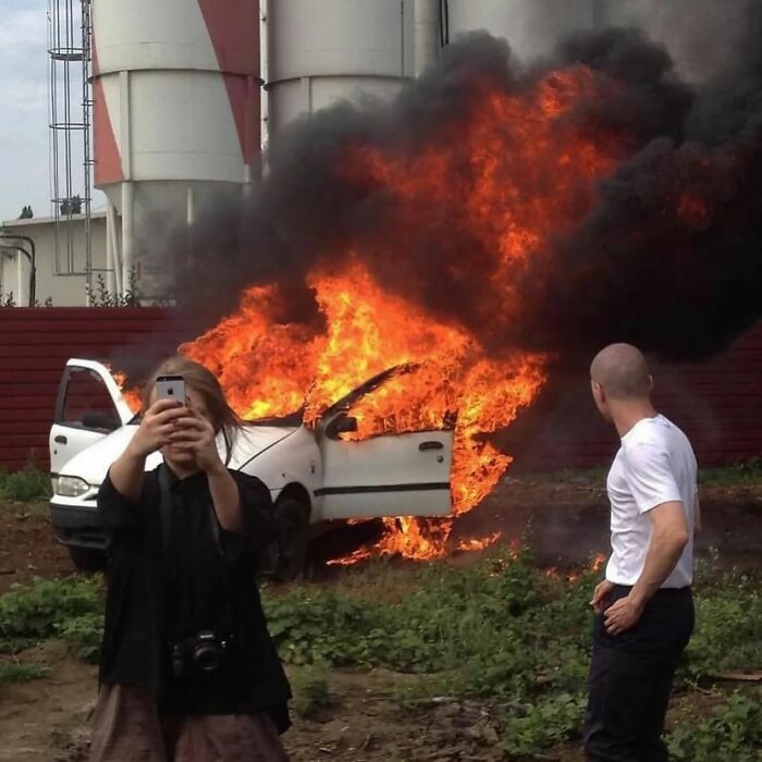 Woman taking selfie with burning car behind her and man watching the fire in a powerful street photo revealing everyday life.