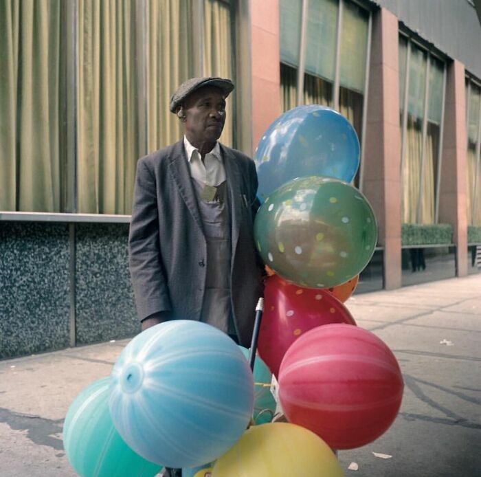 Elderly man holding colorful balloons on city sidewalk in a powerful street photo capturing everyday life moments.