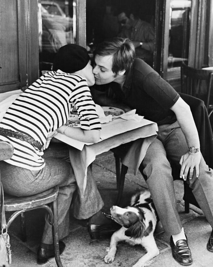 Couple sharing a kiss at an outdoor cafe table with a dog beneath them in a powerful street photo capturing everyday life.
