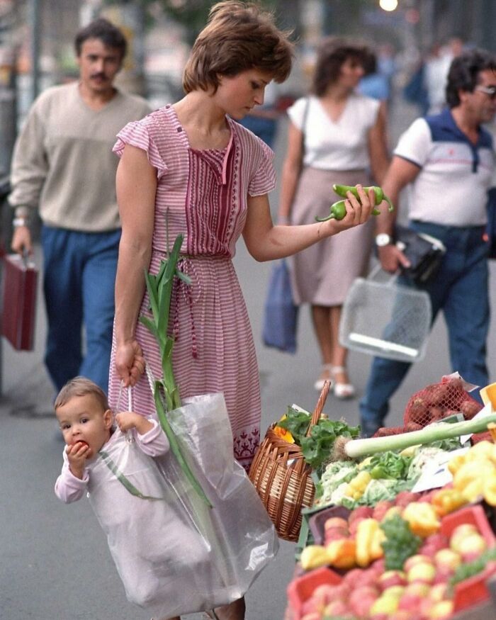 Woman shopping for vegetables at a market, holding a child in a plastic bag, capturing everyday life in a powerful street photo.