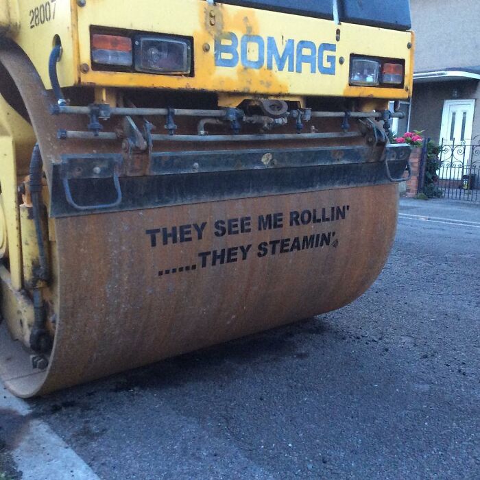 Yellow BOMAG street roller with street art text they see me rollin' they steamin' on its rusty metal drum on asphalt road.