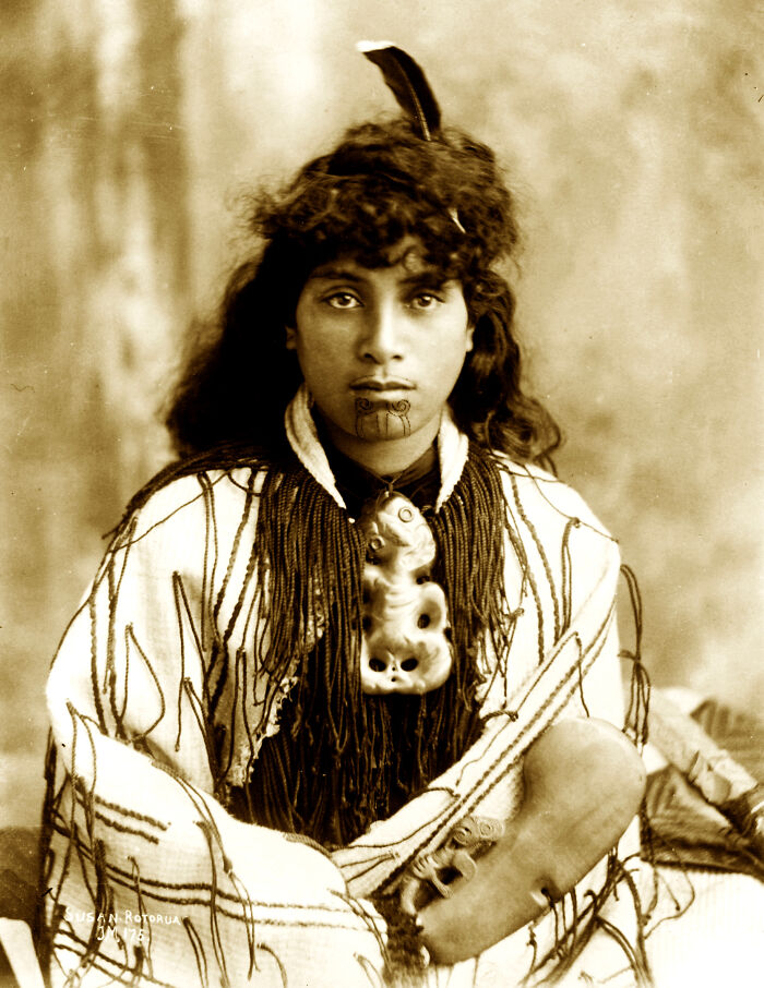 Young Māori woman with traditional Tā moko facial tattoo and carved pendant, dressed in woven Māori cloak from a century ago.