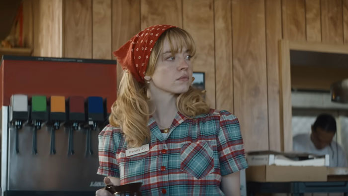 Woman in plaid shirt and red bandana looking aside in rustic diner scene for Sydney Sweeney new movie flop amid backlash.