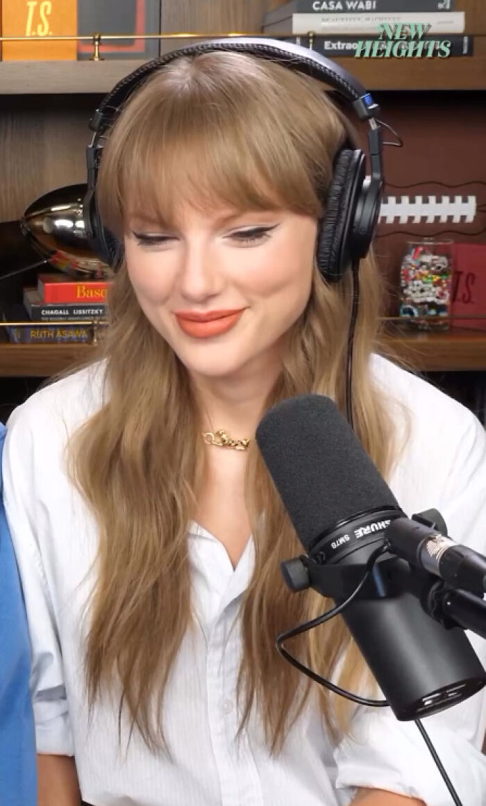Taylor Swift wearing headphones and speaking into a microphone during a podcast recording session in a cozy studio.