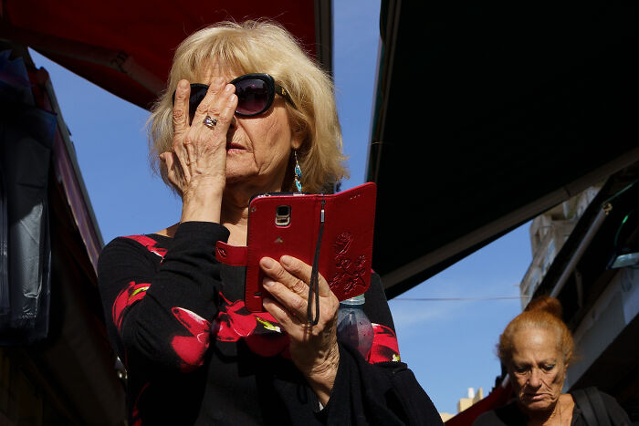 Older woman wearing sunglasses holding red phone case in a street market, showcasing cultural moments captured by photographer.