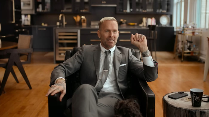 Man in a suit sitting in a chair, discussing the impact of The Biggest Loser and contestants revealing PTSD effects.