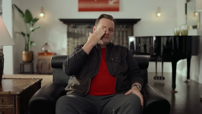 Man sitting on a black leather chair in a living room, wiping his eye, related to Biggest Loser PTSD discussion.