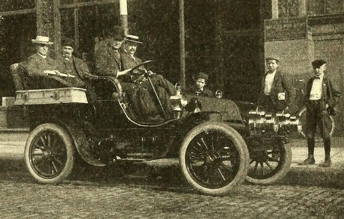 Vintage photo of a wild early car from 100 years ago with passengers and bystanders on a cobblestone street.